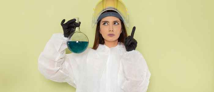 Female chemist in special protective suit holding flask with blue solution and thinking on green surface